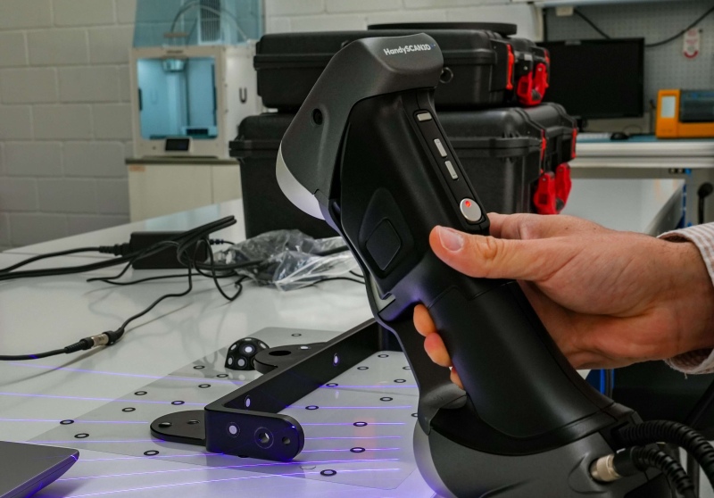 技术人员手持 HandySCAN 3D 三维扫描仪，向计量工作台上的夹具投射蓝色激光线。